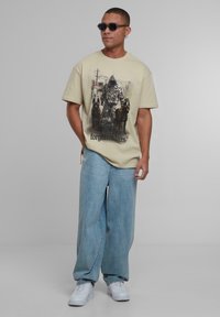 Béžové grafického tričko s centrální ilustrací postavy se psy, spárované s volnými světle modrými džíny a bílými teniskami.
