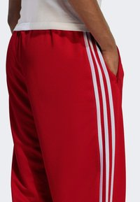 Rote Netz-Sporthosen mit einem weißen Drei-Streifen-Design, ausgestattet mit Seitentaschen und einem elastischen Bund für zusätzlichen Komfort.