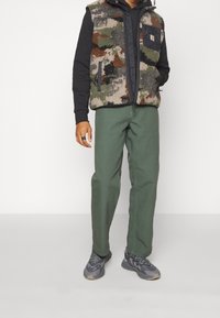 Chaleco de forro polar con un patrón de camuflaje, sobre una sudadera negra, combinado con pantalones verdes y zapatillas grises. Presenta bolsillos laterales y una tela texturizada.