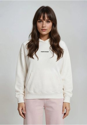 Jeune femme aux longs cheveux bruns porte un sweat à capuche blanc "Pegador" et un pantalon rose clair, debout devant un fond gris clair uni.