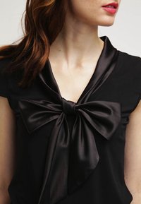 Svart topp med en stor satinrosett vid halsringningen, korta ärmar och en jämn textur. Tyget verkar elegant och strukturerat.