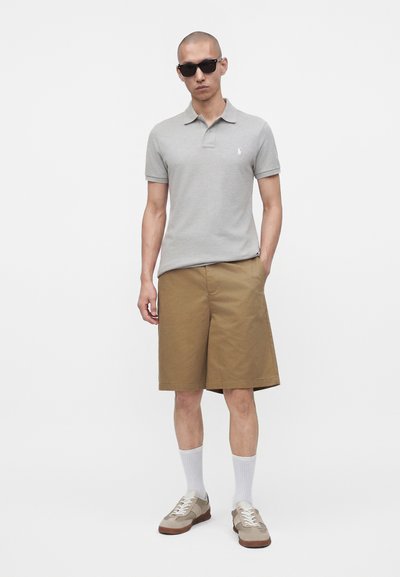 Homme portant des lunettes de soleil noires, un polo gris clair, un short beige longueur genou, des chaussettes blanches et des baskets beiges, debout avec une main dans la poche.