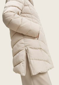 Veste matelassée beige avec des sections matelassées, dotée d'une fermeture éclair sur le côté et de poches. Texture lisse et silhouette décontractée. Idéale pour la chaleur.