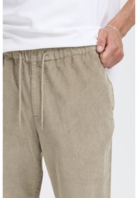 Beige fløjlsbukser med ribbet tekstur, elastisk taljebånd og snørelukning. Vist med en hvid t-shirt.