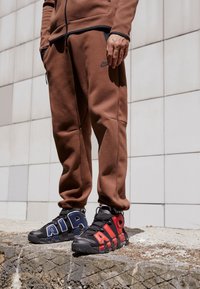 Person som bär bruna Nike träningsbyxor och ojämna Nike Air-sneakers med blå och röda accenter, som står på en betongkant.