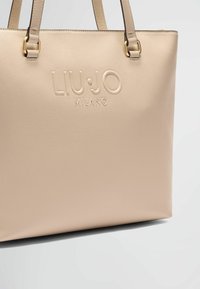 Sac fourre-tout beige en matériau synthétique texturé, arborant un logo embossé "LIU.JO MILANO" et des accents en métal doré.