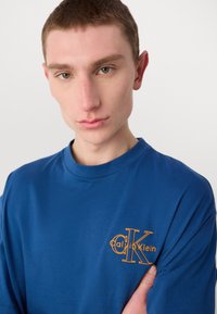 Modrá bavlnená tričko s okrúhlym výstrihom a krátkymi rukávmi, s oranžovým vyšívaným logom Calvin Klein na ľavej hrudi.