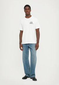 Tricou alb din bumbac cu guler rotund și mâneci scurte, având un logo negru scris de mână pe piept, asortat cu blugi albaștri din denim.