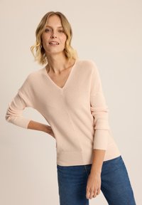 Jasnoróżowy sweter z dekoltem w serek, wykonany z teksturowanego splotu, z prążkowanymi mankietami, w połączeniu z ciemnoniebieskimi dżinsami. Miękka tkanina i luźny krój.