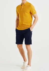 Polo shirt in yellow with textured pattern, three-button placket, paired with navy shorts, rolled cuffs, and white sneakers.