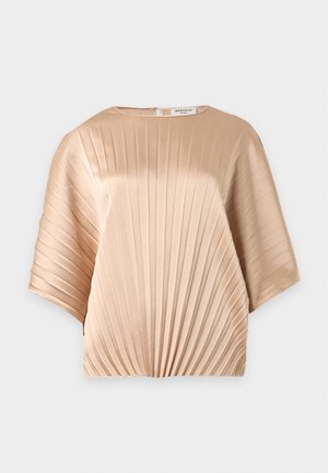 Kobieta ubrana w plisowany beżowy satynowy top, niebieskie dżinsy, czarne szpiczaste buty, trzymająca małą czerwoną cekinową torebkę z łańcuszkiem.