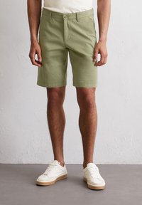Light green cotton shorts with a flat front, two side pockets, and a button closure. Paired with white sneakers featuring beige soles.