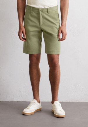 Light green cotton shorts with a flat front, two side pockets, and a button closure. Paired with white sneakers featuring beige soles.