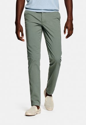 Man wearing light blue shirt, olive green slim trousers, and beige slip-on shoes, standing against plain white background.