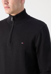 Mand iført sort halv-lynlås sweater med ribbet krave og lille rød, hvid og blå rektangulært logo på brystet.