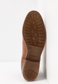 Semelle de chaussure en cuir marron avec motif à losanges et lignes, talon empilé et zone d'étiquette rectangulaire sur fond blanc.