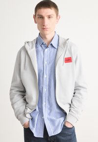 Grå hettejakke med glidelås og lommer foran, med en rød logo-patch, over en lys blå og hvit stripete skjorte med knapper, kombinert med mørke jeans.