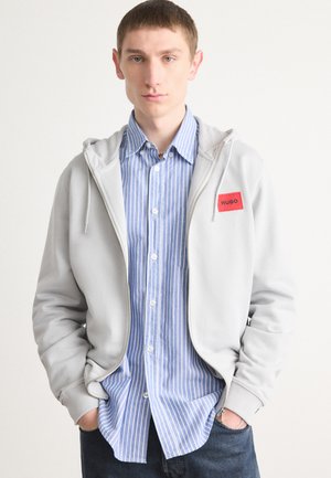 Sudadera gris con cremallera y bolsillo frontal, con un parche de logo rojo, sobre una camisa de botones a rayas azul claro y blanca, combinada con vaqueros oscuros.
