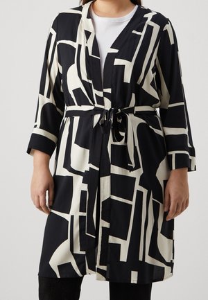 Longue veste à motif géométrique noir et blanc avec ceinture à la taille portée sur un haut blanc et un pantalon noir.
