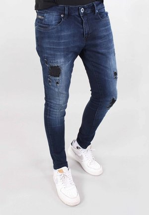 Mand iført mørkeblå, revnede skinny jeans og hvide Nike-sneakers foran en ensartet lys baggrund.