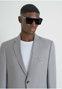 Occhiali da sole neri a forma quadrata, blazer grigio texturizzato, camicia bianca. Il blazer ha un bottone nero singolo e una tasca sul petto. Dettagli del viso chiari.
