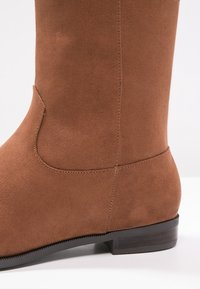 Bottine en daim marron à texture lisse, bout arrondi et petit talon empilé. Présente une couture le long du côté.