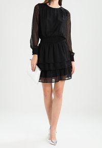 Vrouw in een zwart gemerkte doorzichtige lange mouw jurk, met een witte clutch en zilveren puntige hakken.