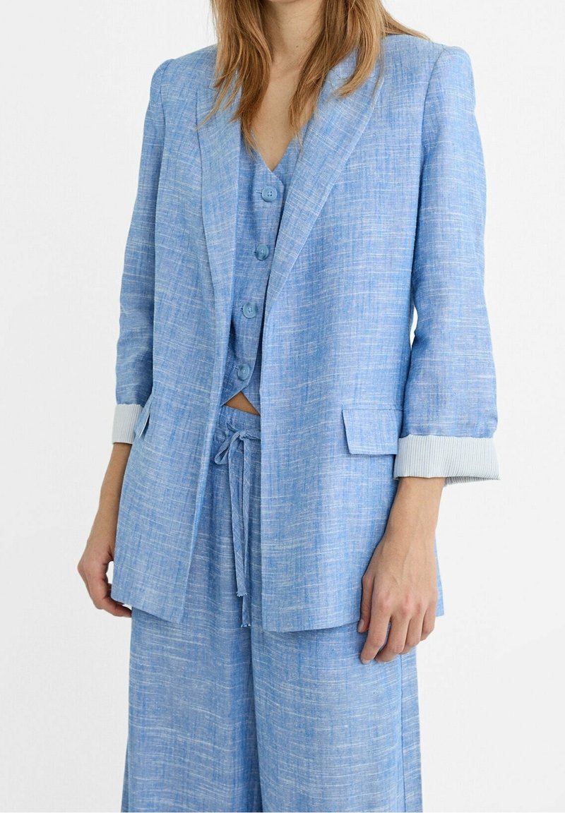 Lichtblauwe linnen blazer met een gestructureerde afwerking, geknipte revers en opgerolde manchetten. Gecombineerd met een bijpassend vest en losse broek.