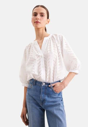 Vrouw draagt een witte eyelet kanten blouse met knopen en pofmouwen, gecombineerd met een blauw hooggetailleerde jeans, hand in de zak, neutrale achtergrond.