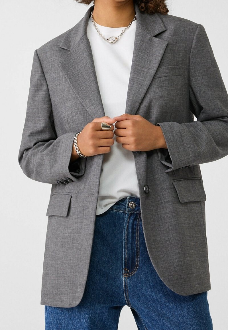 Personne portant un blazer gris par-dessus une chemise blanche et un jean bleu, tenant les bords avant du blazer avec les deux mains.