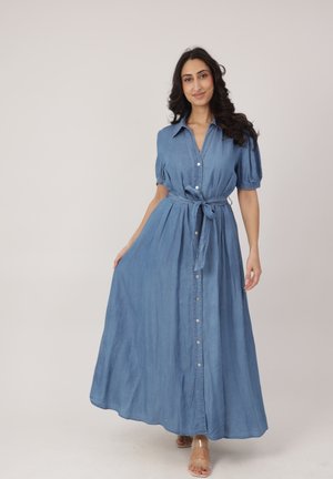 Frau mit langen dunklen Haaren, die ein blaues Maxikleid mit Knöpfen, kurzen Puffärmeln und gebundener Taille trägt, steht vor einem einfarbigen hellen Hintergrund.