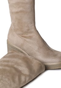Bež suede čizme na visokoj peti sa glatkom teksturom, sa zaobljenim vrhom i elegantnim dizajnom. Jedna čizma djelomično odmotana pored druge.
