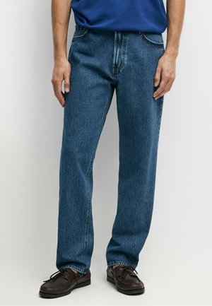 Mand iført blå jeans med lige ben, mørkebrune snørebåndssko og en blå skjorte, stående mod en hvid baggrund.