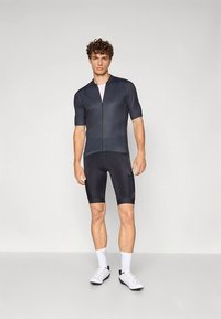 Μπλούζα ποδηλασίας κοντό μανίκι σε σκούρο γκρι, με ύφασμα που απορροφά την υγρασία; συνδυασμένη με μαύρα σορτς ποδηλασίας που διαθέτουν μια λεπτή σχέδιο και τσέπες.