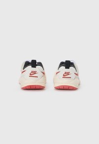 Sneakers Nike z kremową cholewką, czarnymi akcentami i czerwonymi detalami. Posiadają strukturalny materiał, amortyzowaną podeszwę oraz uchwyt z tyłu.