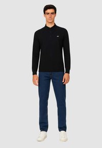 Polo shirt nero a maniche lunghe con chiusura a bottoni e un piccolo logo bianco, abbinato a jeans blu e sneakers bianche, su uno sfondo neutro.