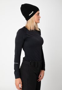 Black beanie with logo, long-sleeve black top with textured accents, and black fitted pants with a belt detail, viewed from the side.