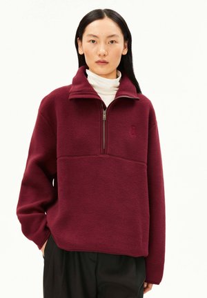 Donna con capelli lisci e neri indossa un pile bordeaux con zip a un quarto sopra un dolcevita bianco, con pantaloni neri, in piedi davanti a uno sfondo neutro.
