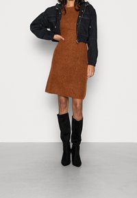 Robe tricotée marron à la texture douce, associée à une veste courte noire et des bottes en daim noir hautes jusqu'au genou. Design simple et coupe ajustée.