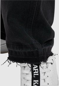 Vaqueros de mezclilla negra desgastados con puños deshilachados, combinados con zapatillas deportivas blancas que tienen una banda de logo negra en la lengüeta.