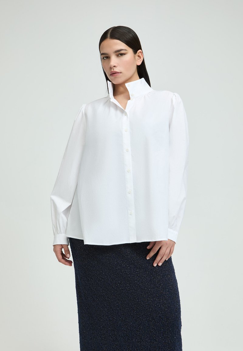White button-up shirt with a high collar, long sleeves, and a loose fit, paired with a textured dark blue skirt.