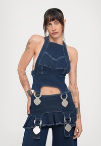 Jeans majica s halter ovratnikom in strukturiranimi žepi, v kombinaciji z razširjeno jeans krilo, ki ima srebrne detajle in volanske dodatke.