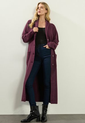 Lang, åben-front cardigan i dyb lilla, lavet af et blødt materiale, med to lommer og store knapper. Sat sammen med mørke skinny jeans og sorte støvler.