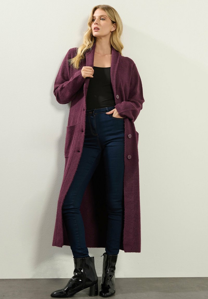 Lång, öppen cardigan i djup lila, tillverkad av ett mjukt material, med två fickor och stora knappar. Tillsammans med mörka tajta jeans och svarta stövlar.