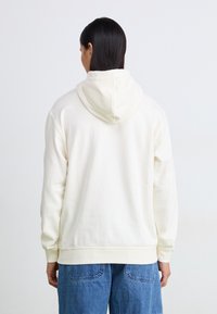 Kremfarget hettegenser med en myk tekstur, med kengurulomme og ribbestrikkede mansjetter, kombinert med blå denimbukser.