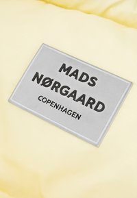 Geel gewatteerde stof met een zilveren label. Het label toont "MADS NØRGAARD" en "COPENHAGEN" in vetgedrukte zwarte letters.