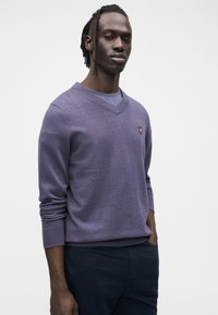 Maglione viola a scollo a V realizzato in tessuto strutturato, con maniche lunghe e piccolo logo giallo sul petto a sinistra. Abbinato a pantaloni scuri.