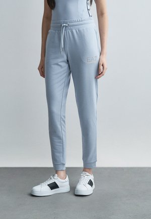 Nő világoskék futónadrágot visel, amelyen az EA7 logó látható a combján, valamint fehér sportcipőt fekete csíkokkal, szürke padlón áll, egyszínű háttér előtt.