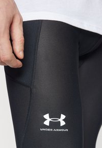 Černé sportovní legíny s přiléhavým střihem a viditelnými švy. Na boku je bílý logo Under Armour. Materiál vypadá hladce a pružně.