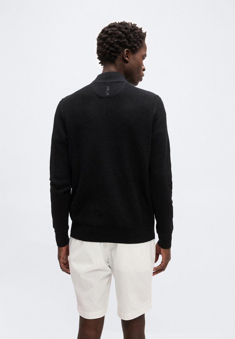 Homme aux cheveux courts et bouclés portant un pull à manches longues noir texturé et un short blanc, debout de dos sur un fond uni.